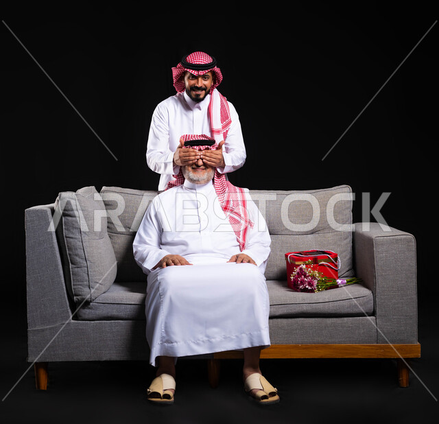 ايماءات السعادة والسرور بعيد ميلاد الوالد، احترام وتقدير الأباء، بورتريه  لشاب عربي خليجي سعودي يرتدي الثوب التقليدي والشماغ يغمض عيني والده بيديه، مفاجأة الأب وإدخال السرور عليه، التربية الصالحة للأبناء، الأعياد ومناسبات عائلية، الطول الكامل للجسم، خلفية سوداء