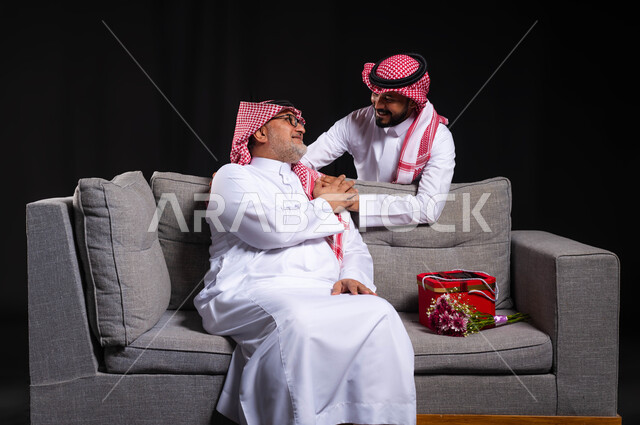 احترام وتقدير الأباء واسعادهم، ايماءات تدل على الإمتنان والرضا والحب، بورتريه لشاب عربي خليجي سعودي يضع يده على كتف والده ويحضر له هدية بمناسبة عيد ميلاده، التربية الصالحة للأبناء، أعياد ومناسبات عائلية سعيدة، ارتداء الأزياء التقليدية السعودية، خلفية سوداء