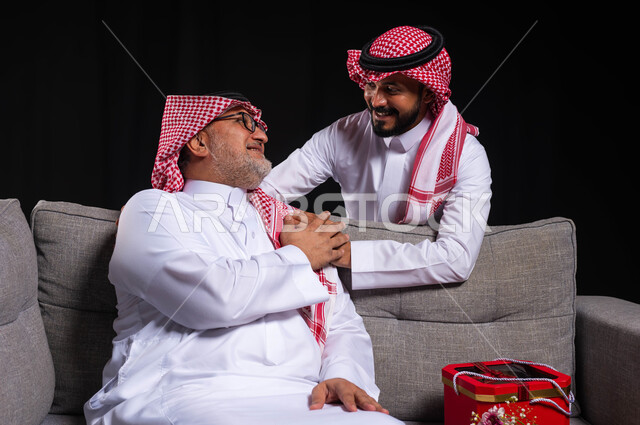 ايماءات تدل على الإمتنان والرضا والحب، بورتريه لشاب عربي خليجي سعودي يضع يده على كتف والده ويحضر له هدية بمناسبة عيد ميلاده، احترام وتقدير الأباء واسعادهم، التربية الصالحة للأبناء، أعياد ومناسبات عائلية سعيدة، ارتداء الأزياء التقليدية السعودية، خلفية سوداء