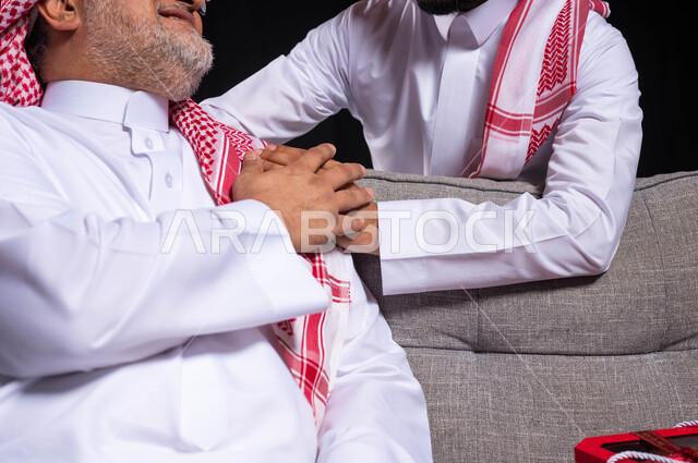 التربية الصالحة للأبناء، احترام وتقدير الأباء واسعادهم، بورتريه مقرب لشاب عربي خليجي سعودي يضع يده على كتف والده بحب حنان، ايماءات تدل على الإمتنان والرضا والحب، أعياد ومناسبات عائلية سعيدة، ارتداء الأزياء التقليدية السعودية، خلفية سوداء