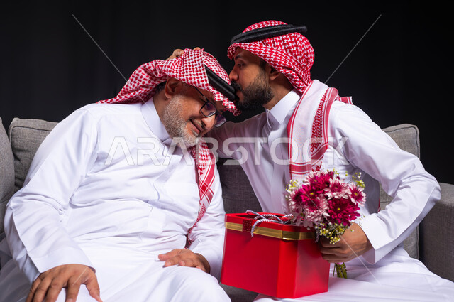 تقدير الآباء والتعبير عن المحبة والألفة، بورتريه لشاب عربي خليجي سعودي يقبل رأس والده ويحمل باقة ورد وصندوق أحمر، تقديم الهدايا للوالد بمناسبة عيد الأب، أعياد ومناسبات ومفاجآت سعيدة، ارتداء الأزياء السعودية التقليدية، خلفية سوداء