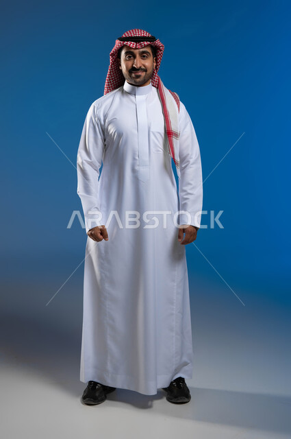 الاهتمام بالمظهر الخارجي، الحفاظ على الصحة الجسدية والنفسية للذكور، مفهوم الرجولة والقوة، بورتريه لشاب عربي خليجي سعودي يرتدي الثوب التقليدي والشماغ يقف باستقامة وينظر إلى الكاميرا بإيماءات الثقة ولاعتزاز بالنفس، صورة لكامل الجسم، خلفية زرقاء