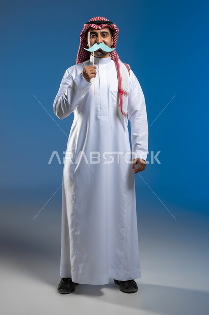 مفهوم الرجولة والقوة، إيماءات البهجة والسرور، بورتريه لشاب عربي خليجي سعودي يرتدي الثوب التقليدي والشماغ يمسك بيده قصاصة ورقية على شكل شارب، ذكرى اليوم العالمي للرجل ١٩نوفمبر، الاهتمام بالصحة الجسدية للذكور، صورة لكامل الجسم، خلفية زرقاء