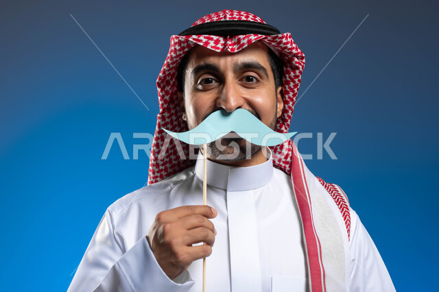 النظر إلى الكاميرا بإيماءات الثقة والاعتزاز بالنفس، مفهوم القوة والرجولة، بورتريه مقرب لشاب عربي خليجي سعودي يرتدي الثوب التقليدي والشماغ يمسك بيده قصاصة ورقية على شكل شارب، ذكرى اليوم العالمي للرجل ١٩نوفمبر، الاهتمام بالصحة الجسدية للذكور، خلفية زرقاء