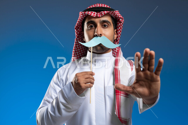 رفع اليد بإيماءات التحذير والتنبيه، ذكرى اليوم العالمي للرجل 19 نوفمبر، الاهتمام بالصحة الجسدية الذكور، بورتريه مقرب لشاب عربي خليجي سعودي يرتدي الثوب التقليدي والشماغ يمسك بيده عود خشبي مع قصاصة ورقية على شكل شارب، خلفية زرقاء