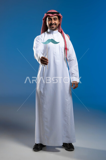 مفهوم الرجولة والقوة، إيماءات البهجة والسرور، بورتريه لشاب عربي خليجي سعودي يرتدي الثوب التقليدي والشماغ يمسك بيده قصاصة ورقية على شكل شارب، ذكرى اليوم العالمي للرجل ١٩نوفمبر، الاهتمام بالصحة الجسدية للذكور، صورة لكامل الجسم، خلفية زرقاء