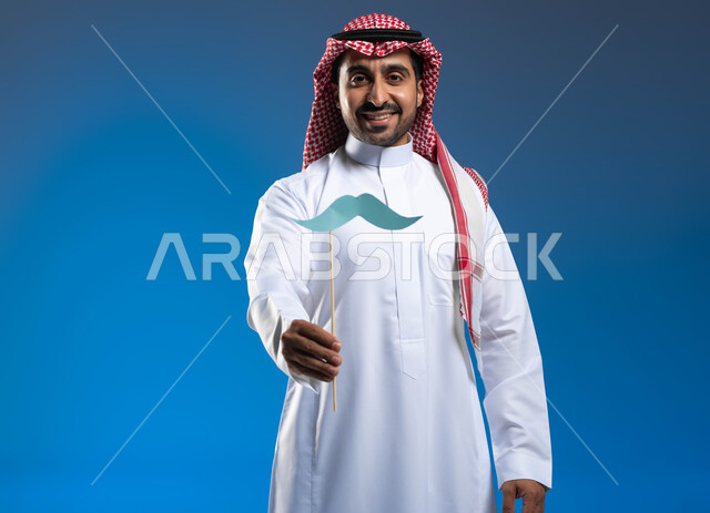 Taking care of the external appearance, looking at the camera with gestures of joy and pleasure, taking care of male health, commemorating November 19, International Men’s Day, a portrait of a Saudi Gulf Arab man wearing traditional clothing holding a wooden stick in his hand with a paper clip in the shape of a mustache, blue background.