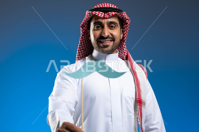 إحياء ذكرى ١٩ نوفمبر اليوم العالمي للرجل، الوقوف باستقامة والنظر إلى الكاميرا بإيماءات البهجة والسرور، الاهتمام بصحة الذكور، بورتريه مقرب لرجل عربي خليجي سعودي يرتدي الزي التقليدي يمسك بيده عود خشبي مع قصاصة ورقية على شكل شارب، خلفية زرقاء