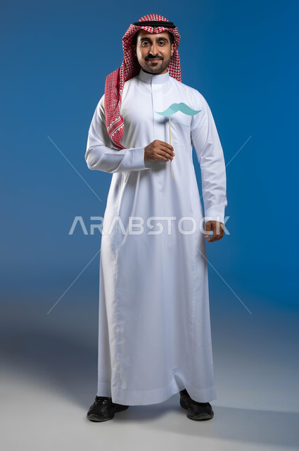 الوقوف باستقامة والنظر إلى الكاميرا بإيماءات الفخر والاعتزاز بالنفس، الاهتمام بصحة الذكور، ذكرى ١٩ نوفمبر اليوم العالمي للرجل، بورتريه لرجل عربي خليجي سعودي يرتدي الزي التقليدي يمسك بيده عود خشبي مع قصاصة ورقية على شكل شارب، صورة لكامل الجسم، خلفية زرقاء