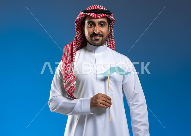 ذكرى اليوم العالمي للرجل، الاهتمام بصحة الذكور، الوقوف باستقامة والنظر إلى الكاميرا بإيماءات الفخر والاعتزاز بالنفس، بورتريه لرجل عربي خليجي سعودي يرتدي الثوب التقليدي والشماغ يمسك بيده عود خشبي مع قصاصة ورقية على شكل شارب، خلفية زرقاء