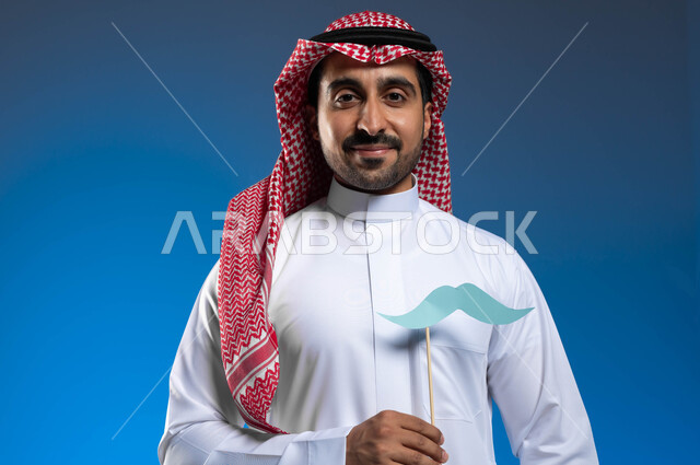 الاهتمام بصحة الذكور، الوقوف باستقامة والنظر إلى الكاميرا بإيماءات الفخر والاعتزاز بالنفس، ذكرى ١٩ نوفمبر اليوم العالمي للرجل، بورتريه مقرب لرجل عربي خليجي سعودي يرتدي الثوب التقليدي والشماغ يمسك بيده عود خشبي مع قصاصة ورقية على شكل شارب، خلفية زرقاء