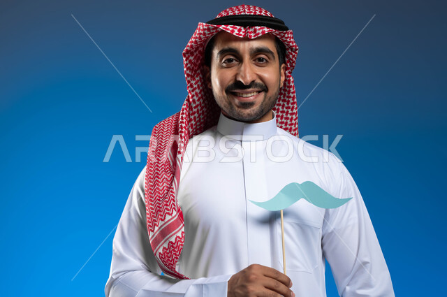 النظر إلى الكاميرا بإيماءات الفخر والاعتزاز بالنفس، ذكرى ١٩ نوفمبر اليوم العالمي للرجل، الاهتمام بصحة الذكور، بورتريه مقرب لرجل عربي خليجي سعودي يرتدي الثوب التقليدي والشماغ يمسك بيده عود خشبي مع قصاصة ورقية على شكل شارب، خلفية زرقاء