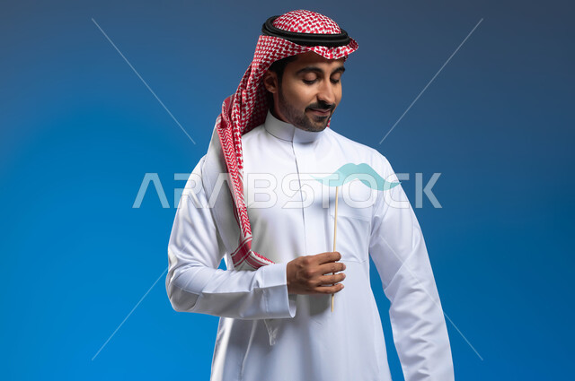 ذكرى ١٩ نوفمبر، الوقوف باستقامة والنظر إلى شعار اليوم العالمي للرجل، بورتريه لشاب عربي خليجي سعودي يرتدي الثوب التقليدي والشماغ يمسك بيده عود خشبي مع قصاصة ورقية على شكل شارب، الاهتمام بالصحة الجسدية والنفسية للذكور، خلفية زرقاء