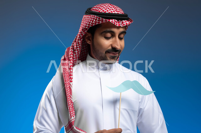 إيماءات الثقة والاعتزاز بالنفس، بورتريه مقرب لشاب عربي خليجي سعودي يرتدي الثوب التقليدي والشماغ يضع في جيبه قصاصة ورقية على شكل شارب يقوم بالنظر إليها، اليوم العالمي للرجل ١٩نوفمبر، الاهتمام بالصحة الجسدية للذكور، خلفية زرقاء