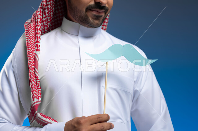 مفهوم الرجولة والقوة، إيماءات الثقة والاعتزاز بالنفس، بورتريه مقرب لشاب عربي خليجي سعودي يرتدي الثوب التقليدي والشماغ يضع في جيبه قصاصة ورقية على شكل شارب، ذكرى اليوم العالمي للرجل ١٩نوفمبر، الاهتمام بالصحة الجسدية للذكور، خلفية زرقاء