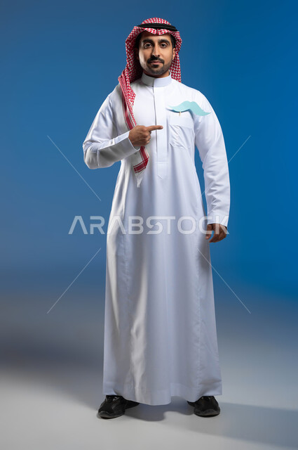 Paying attention to the physical health of males, looking at the camera with gestures of confidence and self-esteem, the concept of strength and masculinity, a portrait of a Saudi Gulf Arab young man wearing the traditional dress and shemagh, putting a piece of paper in the shape of a mustache in his pocket and pointing at it, a full-body photo, blue background.