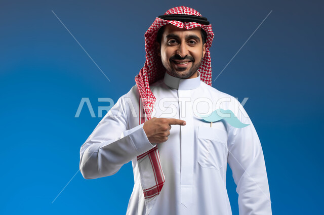 النظر الى الكاميرا بإيماءات الثقة والاعتزاز بالنفس، مفهوم القوة والرجولة، الحفاظ على الصحة الجسدية للذكور، بورتريه لشاب عربي خليجي سعودي يرتدي الثوب التقليدي والشماغ يضع في جيبه قصاصة ورقية على شكل شارب ويقوم بالإشارة اليها، خلفية زرقاء