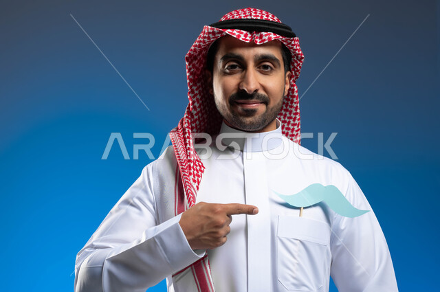 الحفاظ على الصحة الجسدية للذكور، الوقوف باستقامة والنظر الى الكاميرا بإيماءات الثقة والاعتزاز بالنفس، بورتريه مقرب لشاب عربي خليجي سعودي يرتدي الثوب التقليدي والشماغ يضع في جيبه قصاصة ورقية على شكل شارب ويقوم بالإشارة اليها، خلفية زرقاء