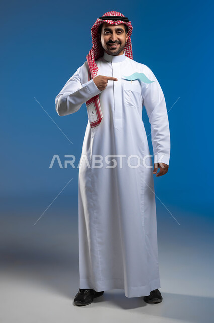 الوقوف باستقامة والنظر الى الكاميرا بإيماءات الثقة والاعتزاز بالنفس، بورتريه لشاب عربي خليجي سعودي يرتدي الثوب التقليدي والشماغ يضع في جيبه قصاصة ورقية على شكل شارب ويقوم بالإشارة اليها، الإهتمام بصحة الذكور، صورة لكامل الجسم، خلفية زرقاء