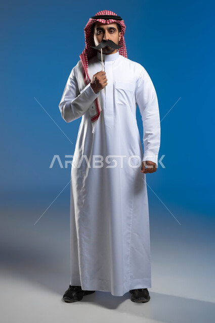 Expressions of firmness and seriousness to alert the need to pay attention to the physical health of males, the anniversary of International Men’s Day, November 19, a portrait of a Saudi Gulf Arab young man wearing the traditional thobe and shemagh, holding a wooden stick in his hand with a paper clip in the shape of a mustache, a full-body portrait, blue background.