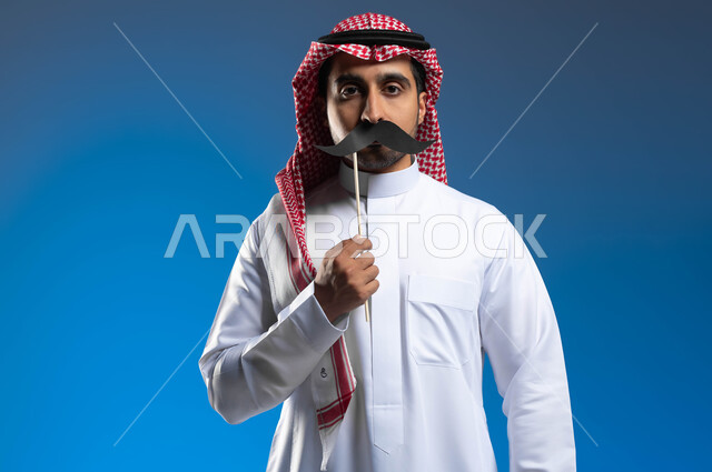 الوقوف باستقامة والنظر إلى الكاميرا بإيماءات الحزم والجدية للتنبيه إلى ضرورة الاهتمام بالصحة الجسدية للذكور، ذكرى اليوم العالمي للرجل 19 نوفمبر، بورتريه لشاب عربي خليجي سعودي يرتدي الزي التقليدي يمسك بيده عود خشبي مع قصاصة ورقية على شكل شارب، خلفية زرقاء