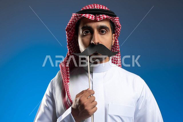 ذكرى اليوم العالمي للرجل 19 نوفمبر، الوقوف باستقامة والنظر إلى الكاميرا بإيماءات الحزم والجدية للتنبيه إلى ضرورة الاهتمام بالصحة الجسدية للذكور، بورتريه مقرب لشاب عربي خليجي سعودي يرتدي الزي التقليدي يمسك بيده عود خشبي مع قصاصة ورقية على شكل شارب، خلفية زرقاء