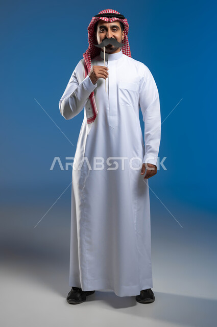 Gestures of confidence and self-esteem, concern for male health, the concept of strength and masculinity, commemoration of International Men’s Day November 19, portrait of a Saudi Gulf Arab young man wearing the traditional thobe and shemagh, holding a wooden stick in his hand with a paper clip in the shape of a mustache, full-body portrait, blue background