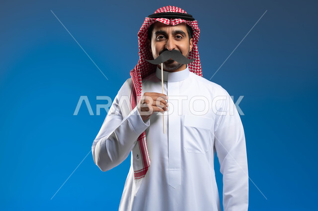 النظر إلى الكاميرا بإيماءات الثقة والاعتزاز بالنفس، الاهتمام بصحة الذكور، مفهوم القوة والرجولة، ذكرى اليوم العالمي للرجل 19 نوفمبر، بورتريه لشاب عربي خليجي سعودي يرتدي الثوب التقليدي والشماغ يمسك بيده عود خشبي مع قصاصة ورقية على شكل شارب، خلفية زرقاء