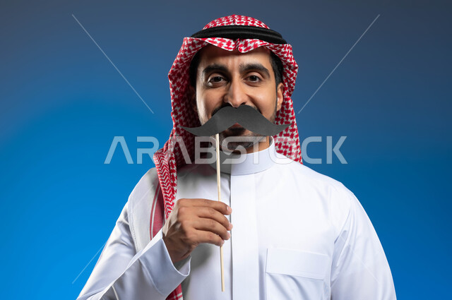 الاهتمام بصحة الذكور، مفهوم القوة والرجولة، النظر إلى الكاميرا بإيماءات الثقة والاعتزاز بالنفس، ذكرى اليوم العالمي للرجل 19 نوفمبر، بورتريه مقرب لشاب عربي خليجي سعودي يرتدي الثوب التقليدي والشماغ يمسك بيده عود خشبي مع قصاصة ورقية على شكل شارب، خلفية زرقاء