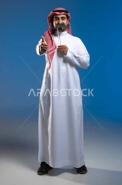 مفهوم القوة والرجولة، ذكرى اليوم العالمي للرجل 19 نوفمبر، رفع اليد بإيماءات الجودة والإعجاب بشيء ما، بورتريه مقرب لشاب عربي خليجي سعودي يرتدي الزي التقليدي يمسك بيده عود خشبي مع قصاصة ورقية على شكل شارب، صورة الجسم بالطول الكامل، خلفية زرقاء