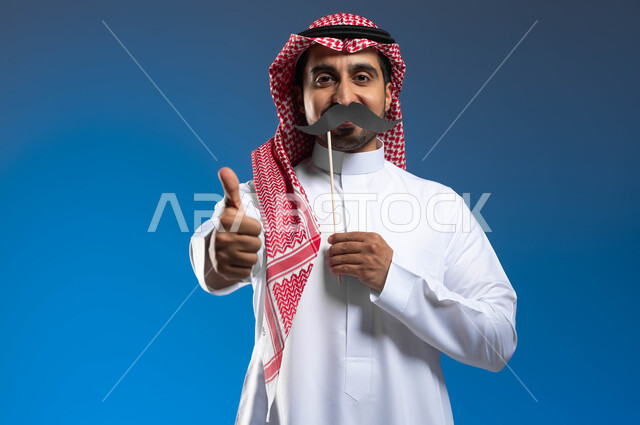 رفع اليد بإيماءات الجودة والإعجاب بشيء ما، مفهوم القوة والرجولة، ذكرى اليوم العالمي للرجل 19 نوفمبر، الاهتمام بصحة الذكور، بورتريه لشاب عربي خليجي سعودي مبتسم يرتدي الثوب التقليدي والشماغ يمسك بيده عود خشبي مع قصاصة ورقية على شكل شارب، خلفية زرقاء