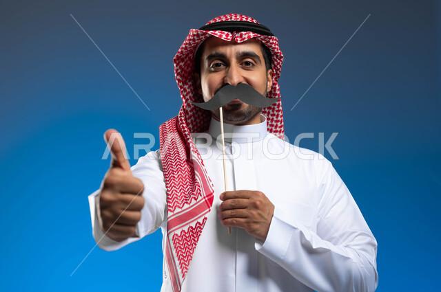 Looking at the camera with expressions of pride and pride, the concept of strength and masculinity, the anniversary of International Men’s Day November 19, raising the hand with gestures of quality and admiration for something, a close-up portrait of a Saudi Gulf Arab young man wearing traditional clothing holding a wooden stick in his hand with a paper clip in the shape of a mustache, blue background