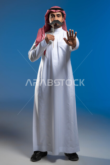 الوقوف باستقامة ورفع اليد بايماءات التحذير والتنبيه، الاحتفال بمناسبة اليوم العالمي للرجل 19 نوفمبر، الاهتمام بصحة الذكور، بورتريه لشاب عربي خليجي سعودي يرتدي الثوب التقليدي والشماغ يمسك بيده عود خشبي مع قصاصة ورقية على شكل شارب، صورة لكامل الجسم، خلفية زرقاء
