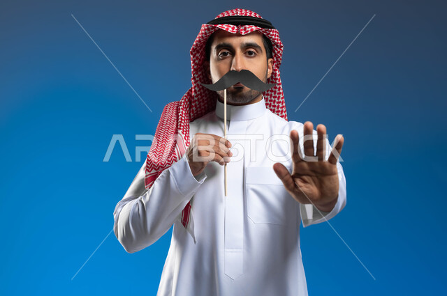 الإمساك بعود خشبي مع قصاصة ورقية على شكل شارب بذكرى اليوم العالمي للرجل 19 نوفمبر، الاهتمام بصحة الذكور، بورتريه لشاب عربي خليجي سعودي يرتدي الثوب التقليدي والشماغ يقوم برفع يده بإيماءات التحذير والتنبيه، خلفية زرقاء