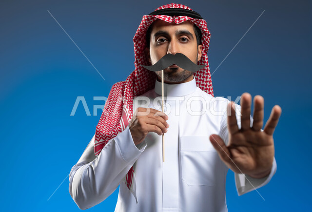 رفع اليد بايماءات التحذير والتنبيه، الاحتفال بمناسبة اليوم العالمي للرجل 19 نوفمبر، الاهتمام بصحة الذكور، بورتريه مقرب لشاب عربي خليجي سعودي يرتدي الثوب التقليدي والشماغ يمسك بيده عود خشبي مع قصاصة ورقية على شكل شارب، خلفية زرقاء