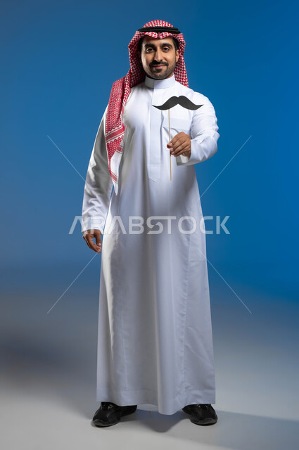 International Men's Day, November 19, the concept of strength and masculinity, gestures of pride and self-esteem, concern for men's health, a portrait of a Saudi Gulf Arab young man wearing the traditional thobe and shemagh, holding a wooden stick in his hand with a paper clip in the shape of a mustache, a full-body portrait, blue background