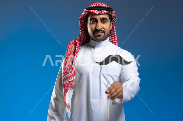 ذكرى19 نوفمبر اليوم العالمي للرجل، مفهوم القوة والرجولة، إيماءات الفخر والإعتزاز بالنفس، الإهتمام بصحة الذكور، بورتريه لشاب عربي خليجي سعودي يرتدي الثوب التقليدي والشماغ يمسك بيده عود خشبي مع قصاصة ورقية على شكل شارب، خلفية زرقاء