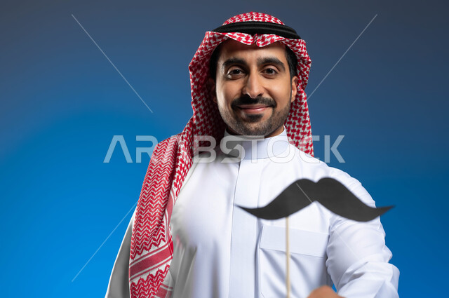 الحفاظ على صحة الذكور، ايماءات الفخر والإعتزاز، النظر الى الكاميرا بإبتسامة والوقوف باستقامة، اليوم العالمي للرجل 19 نوفمبر، بورتريه مقرب لشاب عربي خليجي سعودي يرتدي الثوب التقليدي والشماغ يحمل بيده شعار الشارب، خلفية زرقاء