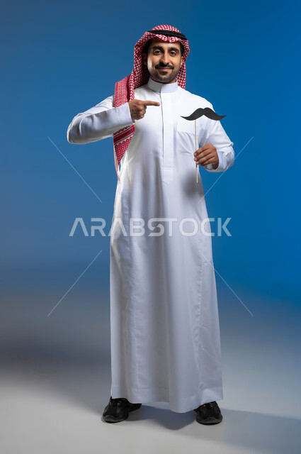 ايماءات الفخر والإعتزاز، النظر الى الكاميرا بإبتسامة والوقوف باستقامة، الإهتمام بصحة الذكور، اليوم العالمي للرجل 19 نوفمبر، بورتريه لشاب عربي خليجي سعودي يرتدي الثوب التقليدي والشماغ يحمل بيده شعار الشارب ويقوم بالإشارة اليه، الطول الكامل للجسم، خلفية زرقاء