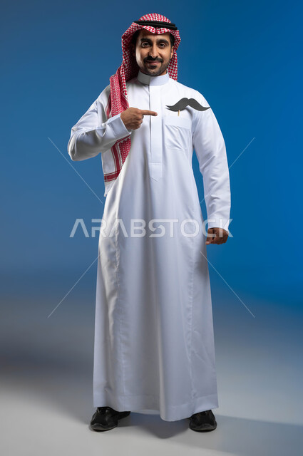 ذكرى اليوم العالمي للرجل، الوقوف باستقامة والنظر الى الكاميرا بإبتسامة، بورتريه لشاب عربي خليجي سعودي يرتدي الثوب التقليدي والشماغ يضع في جيبه شعار الشارب ويقوم بالإشارة اليه، الإهتمام بصحة الذكور، الاحتفال بالمناسبات العالمية، الطول الكامل للجسم، خلفية زرقاء