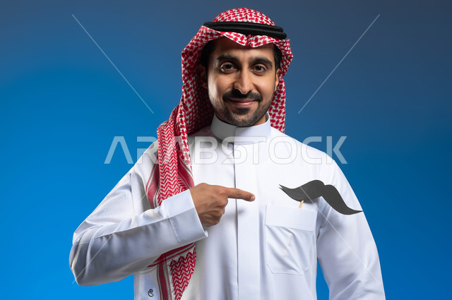 الاحتفال بالمناسبات العالمية، ذكرى اليوم العالمي للرجل، الوقوف باستقامة والنظر الى الكاميرا بإبتسامة، بورتريه مقرب لشاب عربي خليجي سعودي يرتدي الثوب التقليدي والشماغ يضع في جيبه شعار الشارب ويقوم بالإشارة اليه، الإهتمام بصحة الذكور، خلفية زرقاء