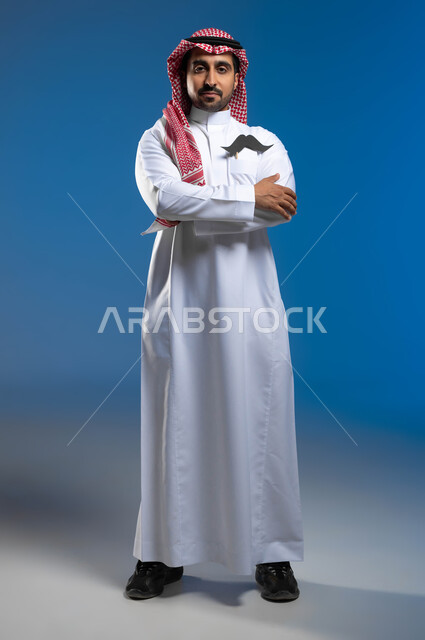 الإحتفال باليوم العالمي للرجل 19 نوفمبر، الحفاظ على صحة الذكور، الإهتمام بمظهر الخارجي،، بورتريه لشاب عربي خليجي سعودي يرتدي الثوب التقليدي والشماغ يضع في جيبيه قصاصة ورقية على شكل شارب، الوقوف باستقامة وأيدي مكتفة، الطول الكامل للجسم، خلفية زرقاء
