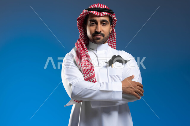 Standing upright with folded hands, celebrating International Men’s Day, November 19, maintaining male health, taking care of external appearance,, close-up portrait of a Saudi Gulf Arab young man wearing the traditional thobe and shemagh, putting a piece of paper in the shape of a mustache in his pockets, blue background.