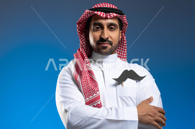 الإهتمام بمظهر الخارجي، الإحتفال باليوم العالمي للرجل 19 نوفمبر، الحفاظ على صحة الذكور، بورتريه مقرب لشاب عربي خليجي سعودي يرتدي الثوب التقليدي والشماغ يضع في جيبيه قصاصة ورقية على شكل شارب، الوقوف باستقامة وأيدي مكتفة، خلفية زرقاء