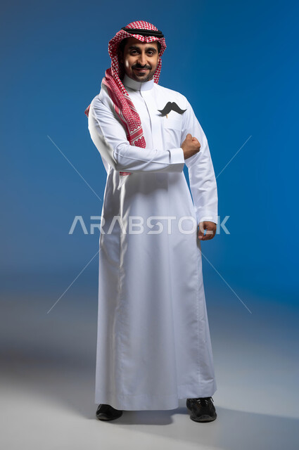 الوقوف باستقامة والنظر الى الكاميرا بإبتسامة، مفهوم الرجولة والقوة، ايماءات قبضة اليد دليل على القوة الجسدية، اليوم العالمي للرجل 19 نوفمبر، بورتريه لشاب عربي خليجي سعودي يرتدي الثوب التقليدي والشماغ يضع في جيبه شعار الشارب، الطول الكامل للجسم، خلفية زرقاء