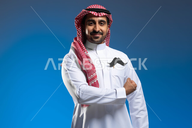 مفهوم الرجولة والقوة، ايماءات قبضة اليد دليل على القوة الجسدية، الإحتفال باليوم العالمي للرجل 19 نوفمبر، بورتريه مقرب لشاب عربي خليجي سعودي يرتدي الثوب التقليدي والشماغ يضع في جيبه شعار الشارب، الوقوف باستقامة والنظر الى الكاميرا بإبتسامة، خلفية زرقاء