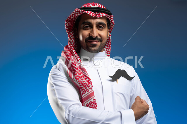 ايماءات قبضة اليد دليل على القوة الجسدية، الإحتفال باليوم العالمي للرجل 19 نوفمبر، مفهوم الرجولة والقوة، بورتريه مقرب لشاب عربي خليجي سعودي يرتدي الثوب التقليدي والشماغ يضع في جيبه شعار الشارب، الوقوف باستقامة والنظر الى الكاميرا بإبتسامة، خلفية زرقاء