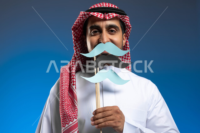 الحفاظ على صحة الذكور، الإهتمام بمظهر الخارجي، الاحتفال بالمناسبات العالمية، اليوم العالمي للرجل 19 نوفمبر، بورتريه مقرب لشاب عربي خليجي سعودي يرتدي الثوب التقليدي والشماغ يحمل عود خشبي عليه قصاصات ورقية على شكل شارب، خلفية زرقاء