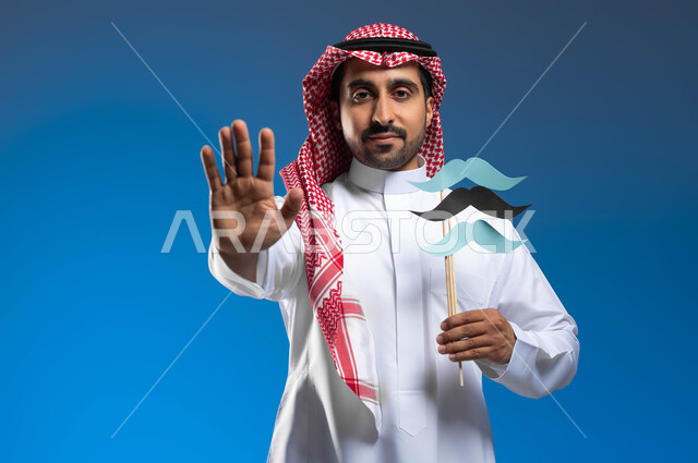 الإهتمام بصحة الذكور، رفع اليد بايماءات التحذير والتنبيه، الاحتفال بمناسبة اليوم العالمي للرجل 19 نوفمبر، بورتريه مقرب لشاب عربي خليجي سعودي يرتدي الثوب التقليدي والشماغ يحمل عود خشبي عليه قصاصات ورقية على شكل شارب، خلفية زرقاء