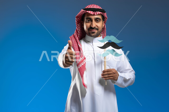 رفع اليد بايماءات الجودة والإعجاب بشيء ما، الفخر والإعتزاز بالنفس، ذكرى اليوم العالمي للرجل 19 نوفمبر، مفهوم الإهتمام بصحة الرجال، بورتريه مقرب لشاب عربي خليجي سعودي يرتدي الثوب التقليدي والشماغ يحمل عود خشبي عليه قصاصات ورقية على شكل شارب، خلفية زرقاء